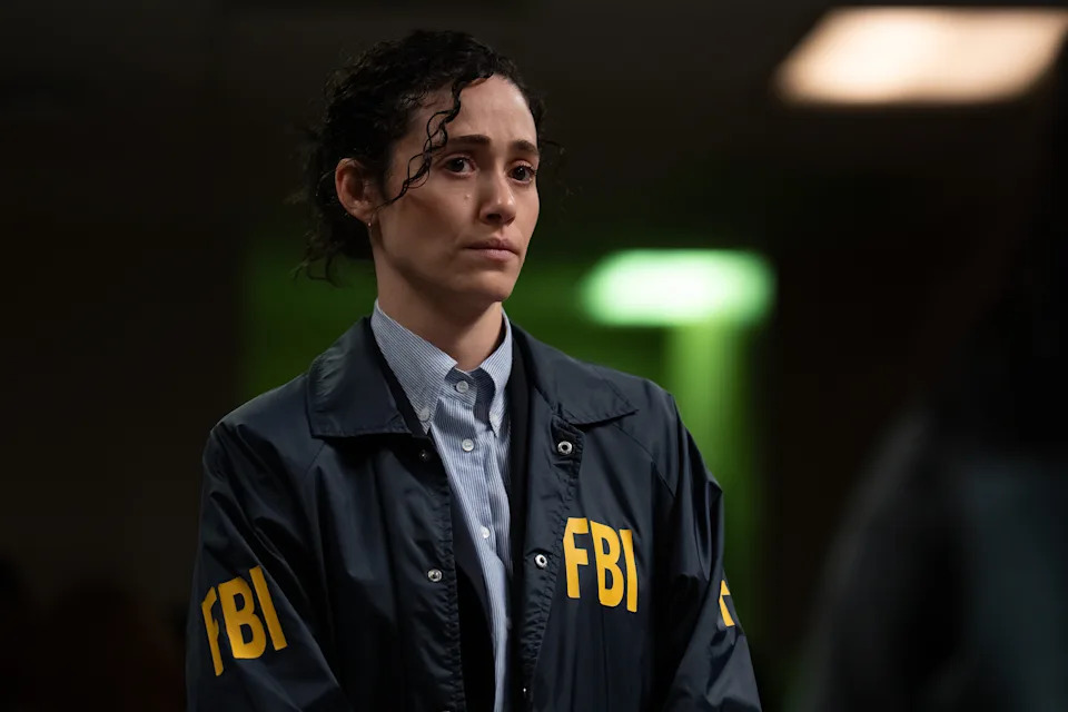 Emmy Rossum appears in a scene from Furious, wearing a navy FBI windbreaker with large yellow "FBI" lettering over a light blue button-down shirt. Her dark curly hair is pulled back with loose strands framing her face, and she looks ahead with a solemn, emotional expression. The dimly lit background is softly blurred, drawing focus to her as she stands during what appears to be a tense moment.