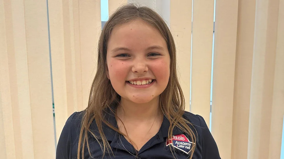 A young girl with long light-brown hair. She is wearing a dark blue and red top with her school's logo on it. She is stood in front of some cream-coloured window blinds.