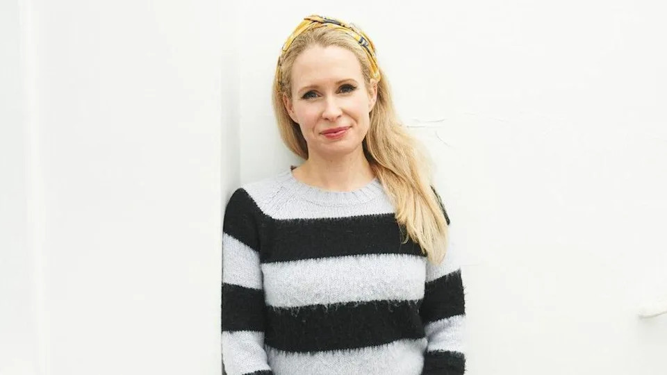 Lucy Beaumont - a woman with long blonde hair and a yellow headband - is leaning against a wall and smiling. She is wearing a white and black striped jumper.