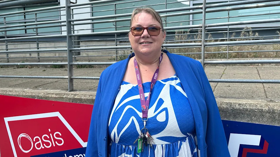 A woman with tied-back light hair, wearing sunglasses. She is wearing a light blue jacket over a white and blue patterned dress, and has a purple lanyard. She is stood outside in front of a red and blue sign with 'Oasis Academy' written on it.