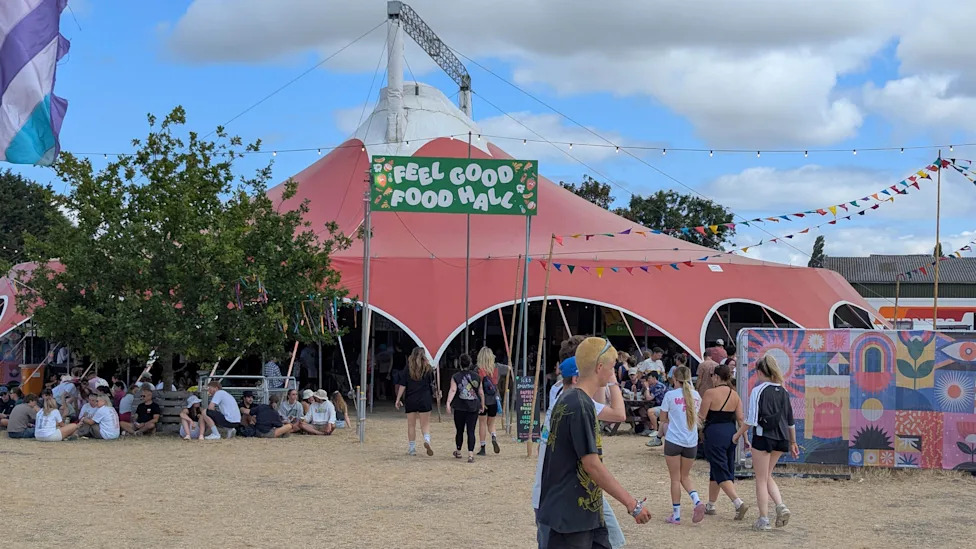 People walking by the Feel Good Food Hall tent at Truck Festival. There is a group sitting beside it. There is a colourful mural on the right. It is cloudy.