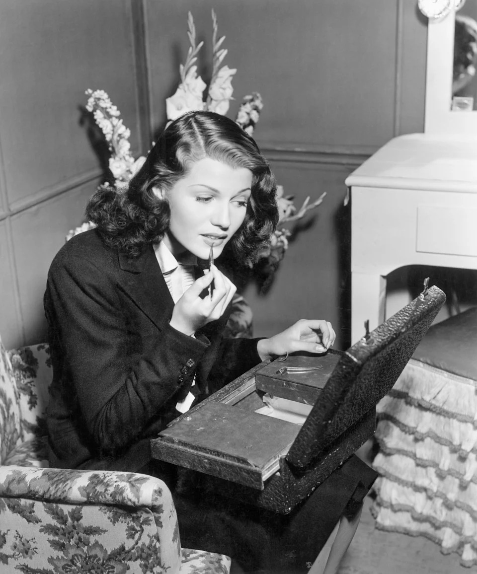 Rita Hayworth in vintage clothing applies lipstick while seated at a vanity with flowers.