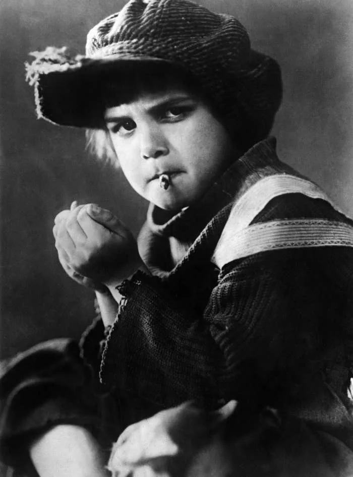 Jackie Coogan dressed in early 20th-century street clothes, wearing a cap, looks intently at the camera, holding a small object to their mouth