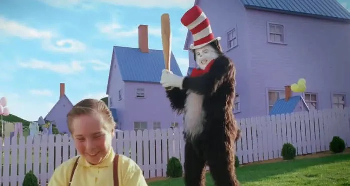Person in a cat costume with a red and white striped hat playfully holds a bat behind a smiling child in a yard