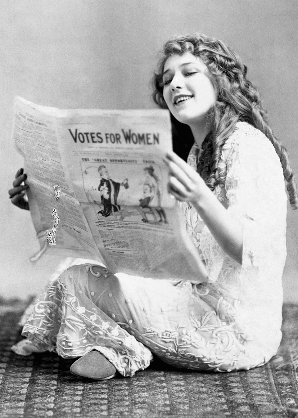 Mary Pickford in a long dress sits on a patterned rug, smiling while reading a newspaper with the headline "Votes for Women."