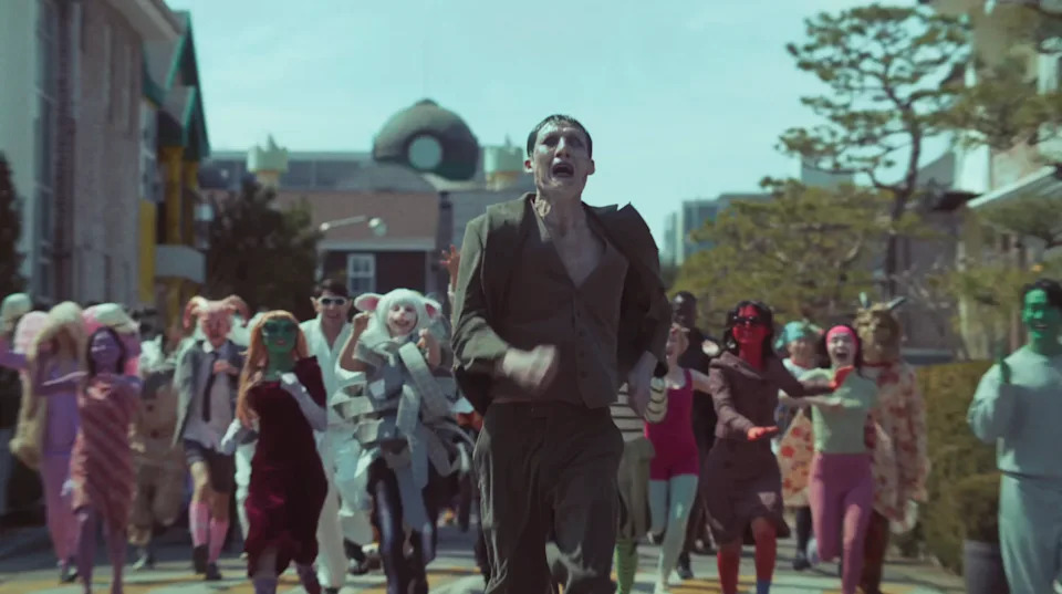 People in colorful costumes chase a panicked person down a street, creating a lively and surreal scene