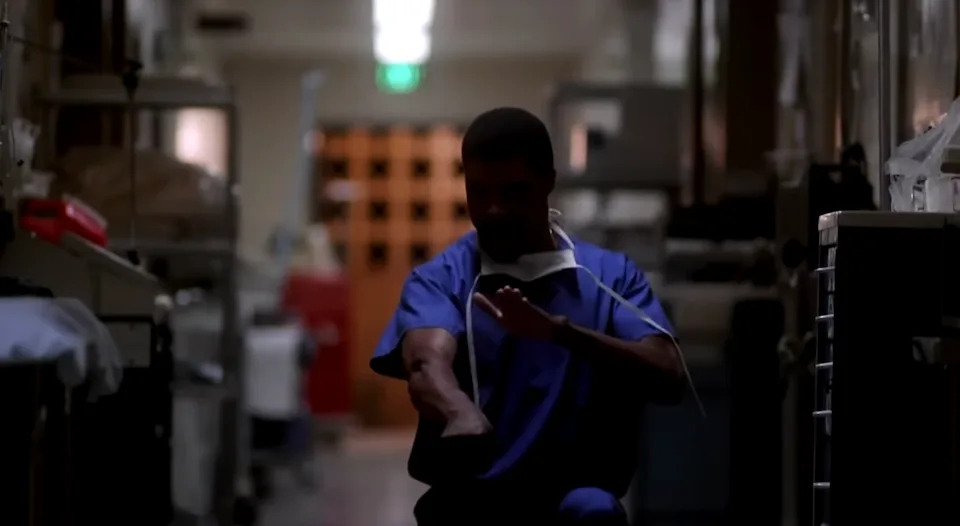 A person in medical scrubs performs a hand-washing gesture in a dimly lit corridor