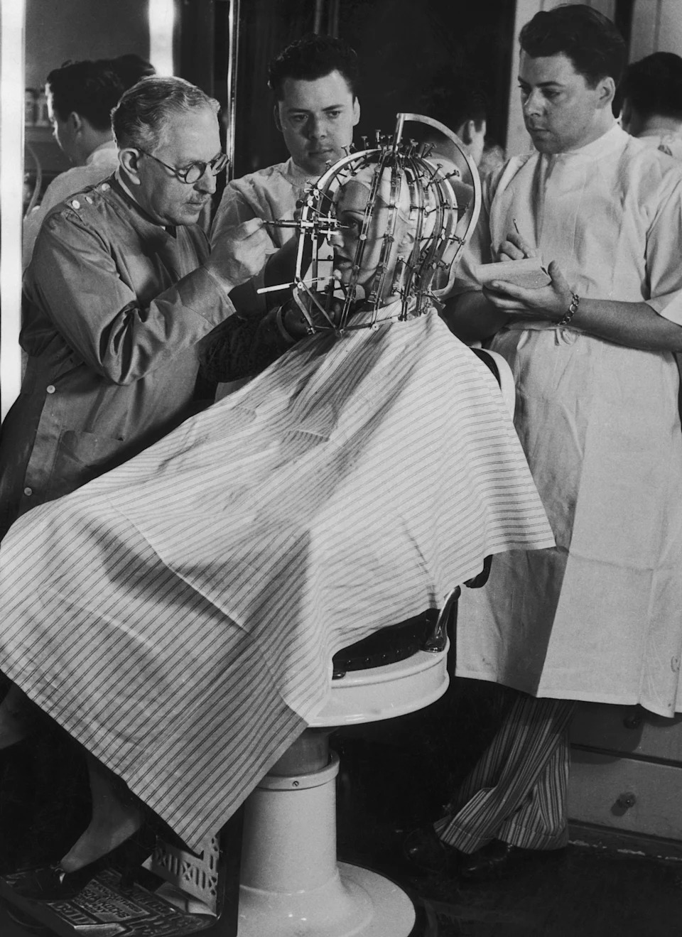 Max Factor and two other men work on a woman seated with her head in a complex metal hair-waving machine at a salon
