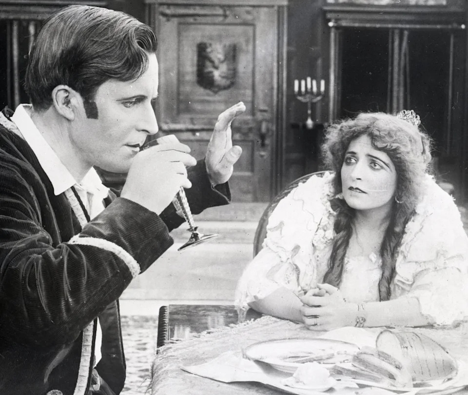 William D Taylor, dressed formally, drinks from a glass while a woman in a period gown looks on with a concerned expression, seated at a table