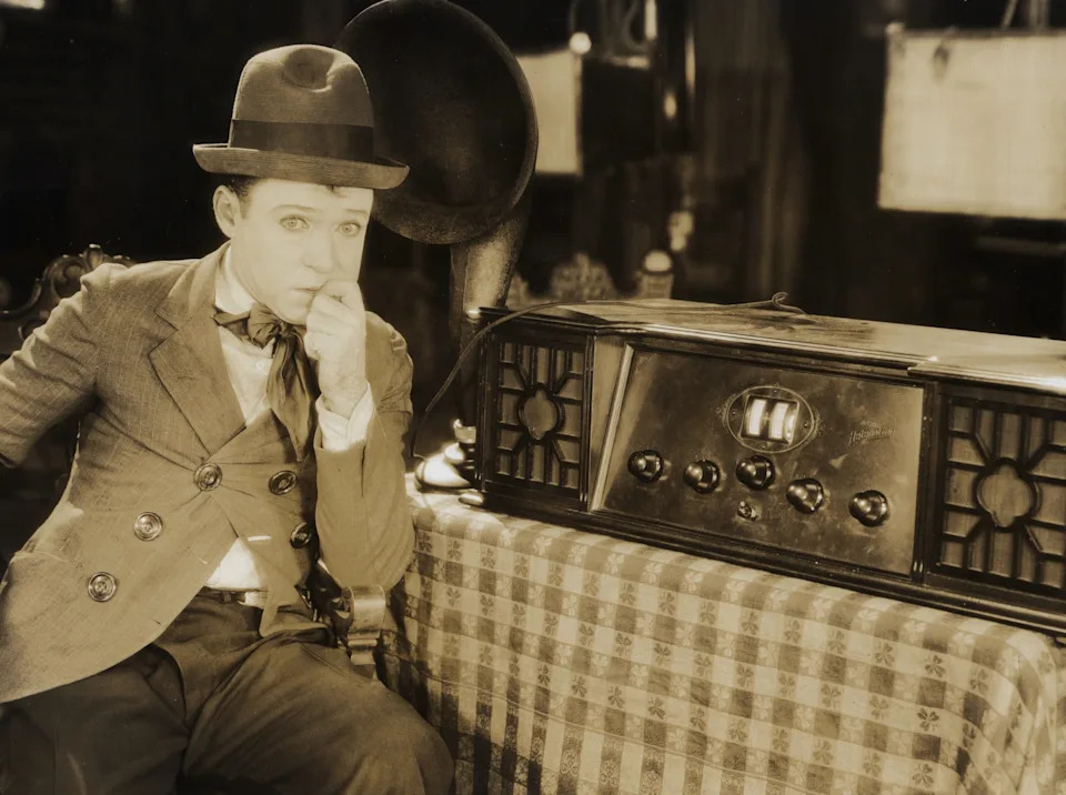 Harry Langdon in a vintage suit and bowler hat sits thoughtfully by an old-fashioned radio on a table