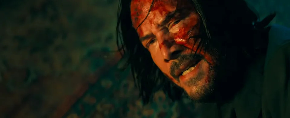 A disheveled man with long hair has a bloodied face and a grim expression, suggesting a tense or dramatic scene