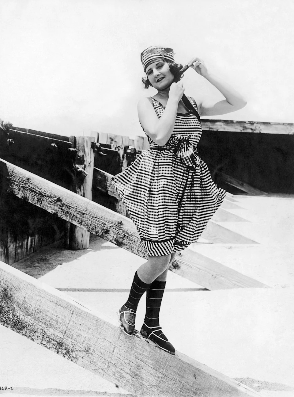 Vintage photo of Vera Stedman in a striped bathing suit, matching cap, and striped socks, posing playfully on a wooden structure by the beach