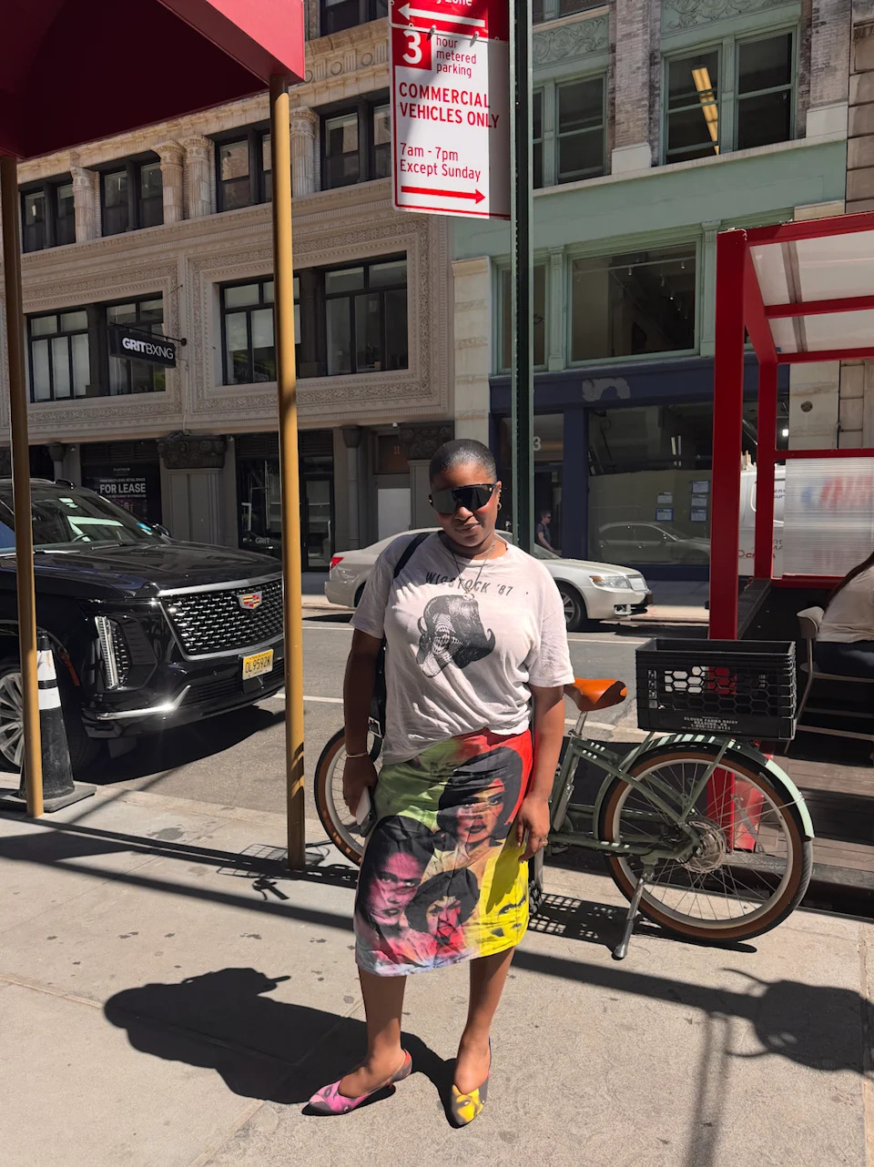 A person wearing a graphic t-shirt and colorful skirt stands on the sidewalk near parked cars and a bicycle.