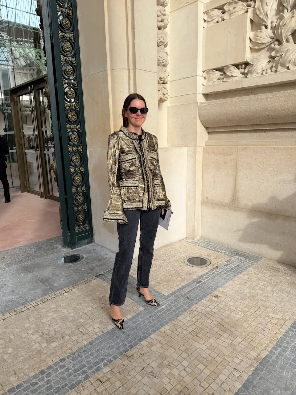 Woman in a stylish gold jacket and jeans stands outside an ornate building, smiling with sunglasses on.