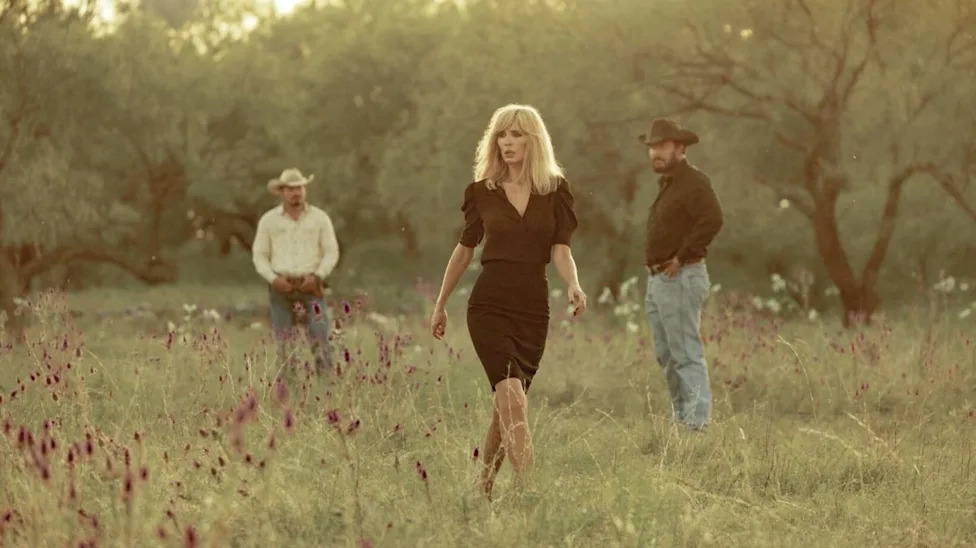 A woman in a black dress walks through a field with two men in the background, surrounded by wildflowers and trees.