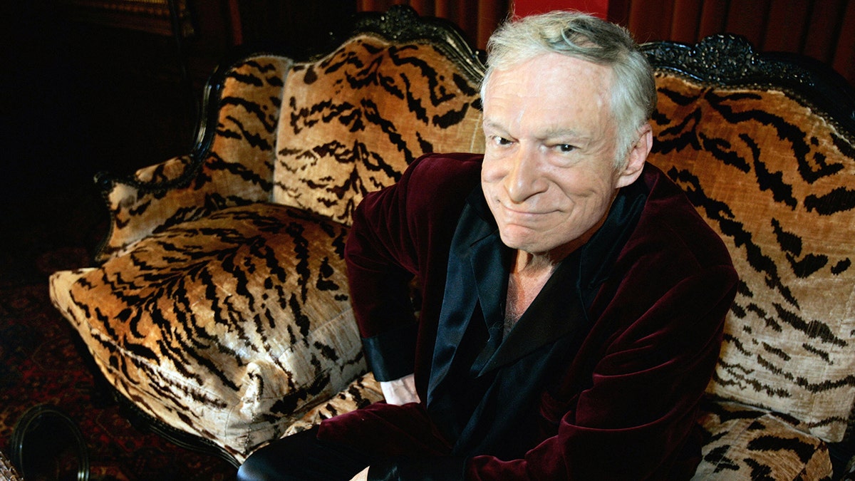 Hugh Hefner in his signature maroon velvet robe soft smiles and looks up sitting on a tiger print couch