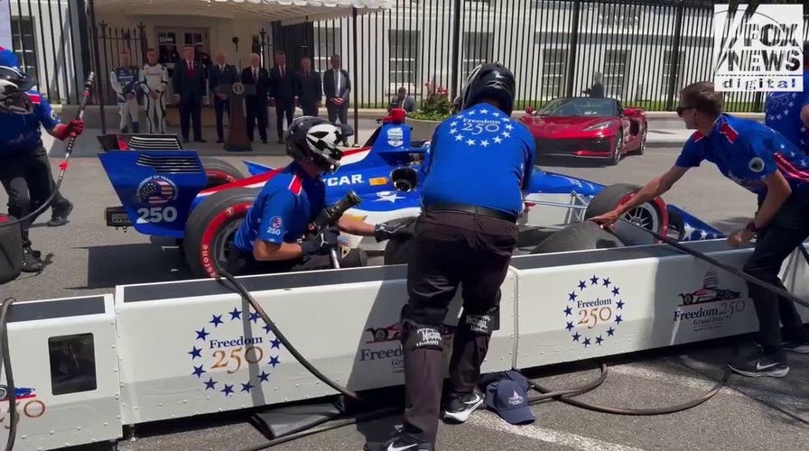 An INDYCAR driver zooms towards pit crew at preview of America 250 Grand Prix.