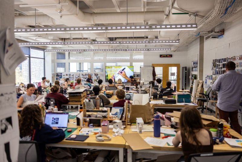 Students participate in a studio workshop, listening to a presentation while surrounded by prototypes, sketches, and design concepts in progress.