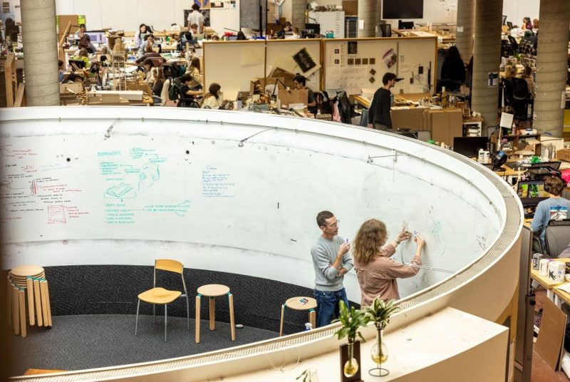 Industrial design students work throughout a studio space while two people collaborate on ideas inside the Kiva, a circular brainstorming area surrounded by whiteboards.