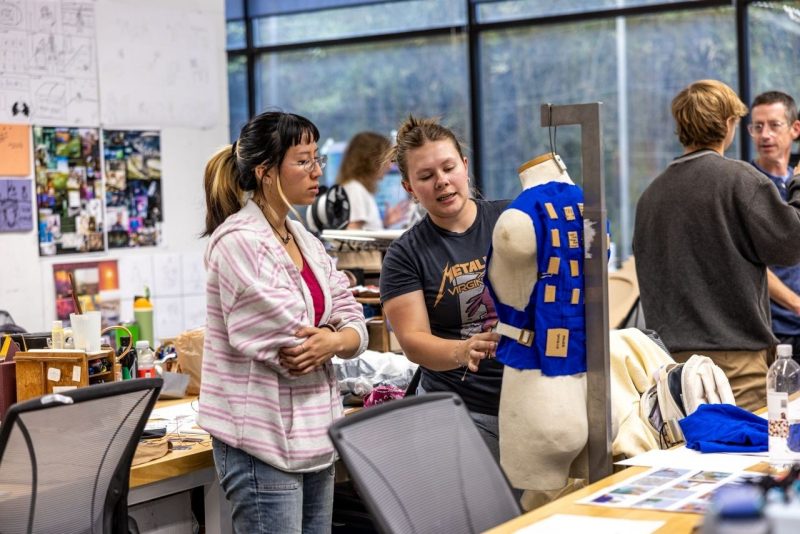 Two students examine and discuss a wearable prototype mounted on a dress form in the design studio.