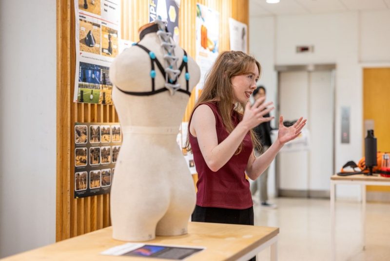A student presents her speculative design project during a final exhibition, standing beside a prototype wearable device displayed on a dress form.