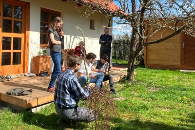 A group of people sit on a deck on a sunny day braiding willow branches together into short whips