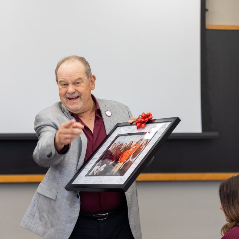 A man in a suit smiles and points to the camera with one hand. In the other, he holds a framed photo with a red bow on it.