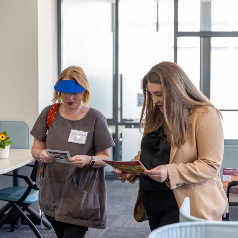 Two women stand together in a well-lit room, both examining papers in their hands.