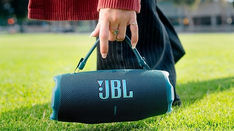 A person picking up the JBL Charge 6 Bluetooth speaker from the grassy ground.