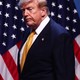 Photograph of Donald Trump frowning. Four American flags are behind him.