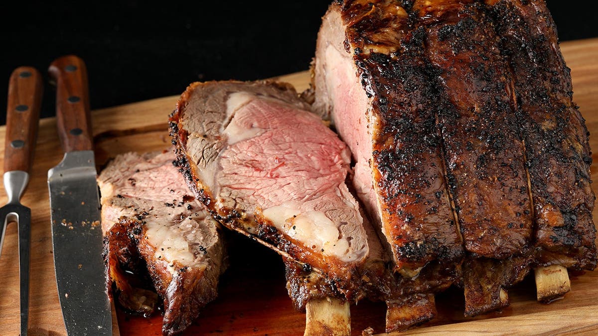 A cooked and sliced piece of prime rib sits on a cutting board with a knife and fork.