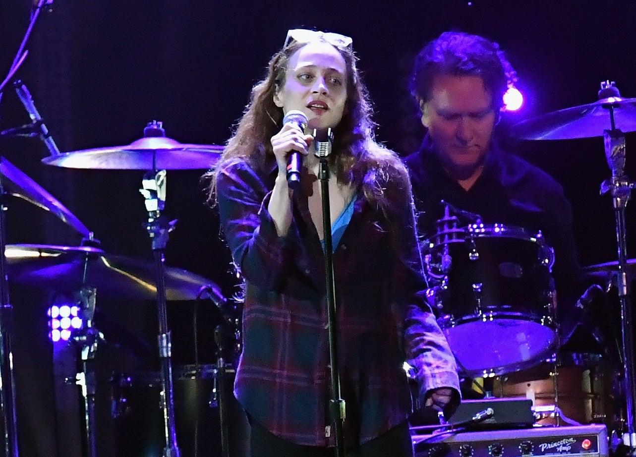 Fiona Apple on stage holding a microphone, wearing a plaid shirt, with a drummer playing in the background
