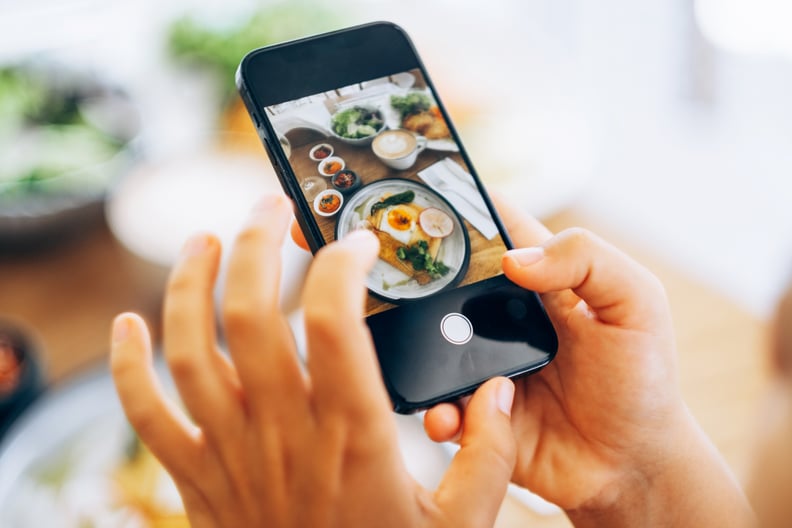Hands holding smartphone taking a photo of delicious plated breakfast food with coffee and sides on a bright table