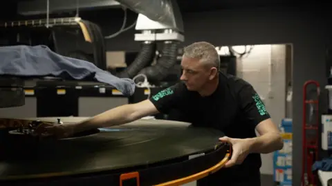 A man with silver hair and black T-shirt leans across an enormous reel of film