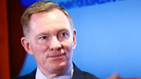 Getty Images A man with short fair haired wears a black suit and blue shirt. He stands in front of a blue screen.