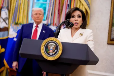 Getty Images Jeanine Pirro is in the Oval Office speaking from a podium bearing the presidential crest. She is wearing a white stui and a gold brooch on her left lapel. Donald Trump is standing behind her, slightly out of focus, wearing his trademark royal blue suit and red tie.