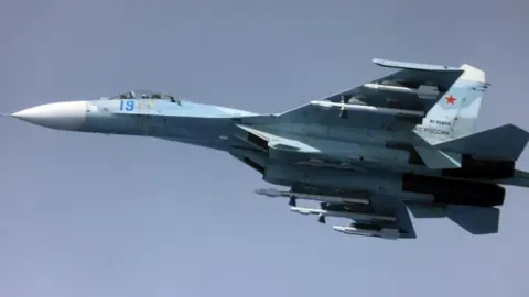 UK Ministry of Defence Image of a Russian Su-27, a whit and blue coloured fighter jet, airborne against a clear blue sky.