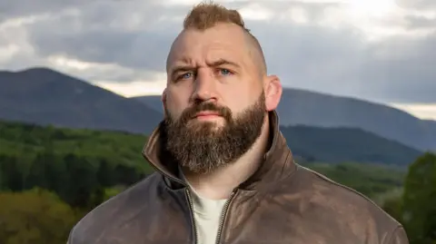 BBC Joe Marler pictured wearing a jacket and looking stern with trees and mountains in the background