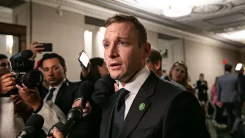 Bloomberg via Getty Images Max Miller, wearing a suit and tie, speaking to reporters holding up microphones in a capitol building