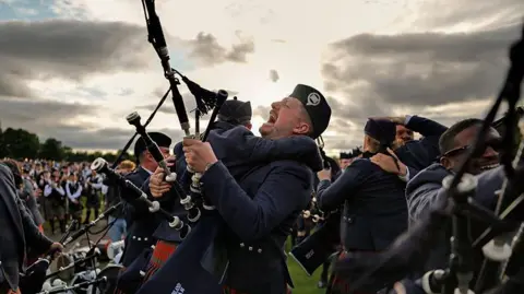 Getty Images Two pipe band members in matching dark blue uniforms and red-and-green tartan kilts are embracing. One of them is holding bagpipes and his mouth is open in joy. There are other pipers hugging each other around them. There is low sun behind the clouds creates strong backlighting.