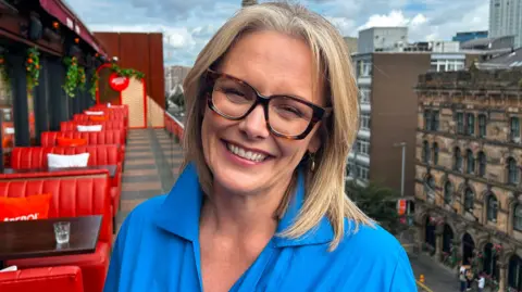 A woman in a blue shirt with blonde hair and glasses. She is standing on the balcony of a building with red sofas behind her. The sky is blue with grey clouds.