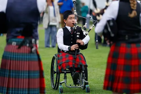 Getty Images Young person in a wheelchair playing a set of bagpipes. They are wearing a traditional piper outfit with hat