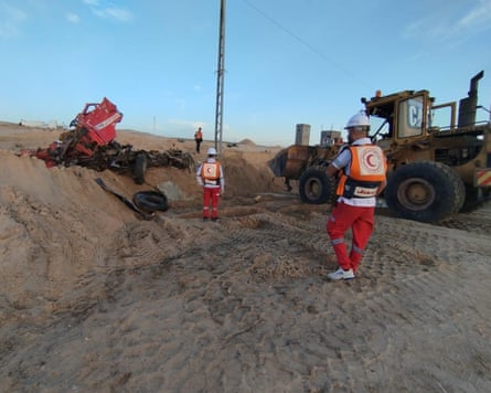 Two Red Crescent paramedics standing by a mound of bulldozed sand with the ruins of vehicle protruding