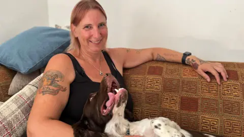 BBC A woman with tattoos sitting on a brown sofa with her springer spaniel. She is looking ahead and smiling and her spaniel is looking up at her with her mouth open,