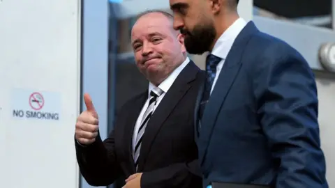 ABC News/Jamie McKinnell A man in a black suit and tie, giving a thumbs up to the camera, with another mad standing by his side