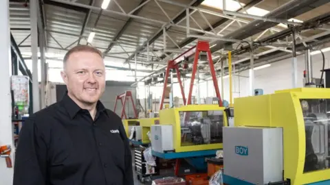 Clarity Plastics Head and shoulders of a man, smiling, wearing a black shirt, standing in a factory with a row of yellow manufacturing units in the background