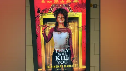 PA Media A film poster on an underground station wall shows a blood-splattered woman standing in front of an ornate doorway. She wears a blood-stained shirt, has blood splattered across her face, and holds a weapon resting over her shoulder. Red blood splatters cover the background behind her. The text reads: "From the producers of They Will Kill You. In cinemas March 27."