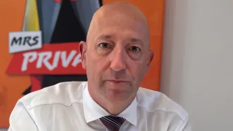 BBC Paul Vane sitting at a desk with his hands clasped. He has a serious expression, and is wearing a white shirt and stripey tie. He is bald. There are campaign posters visible in the background.