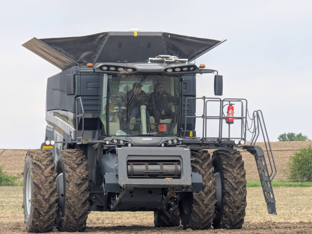 Two men ride in the cab of a tractor in a field.