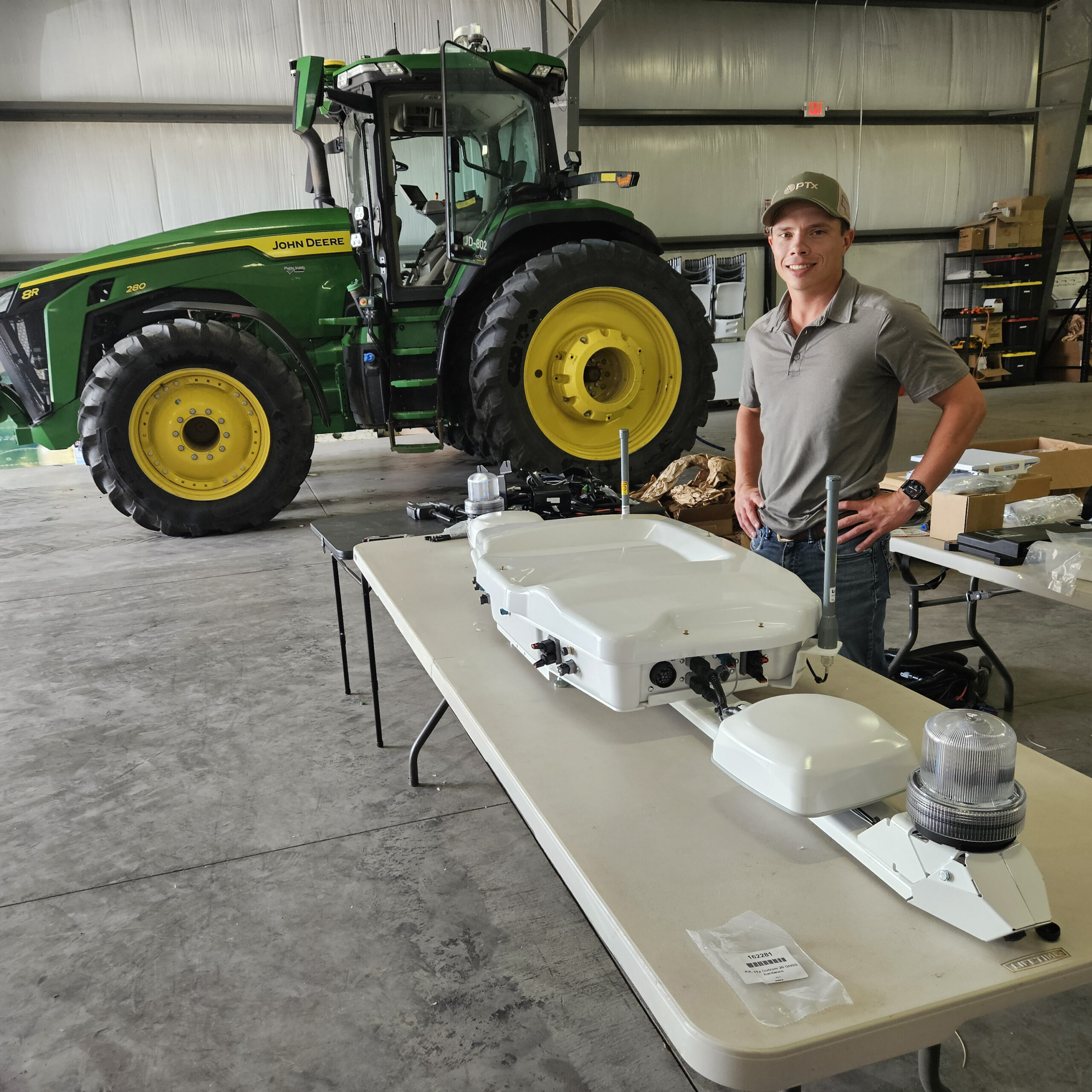 Josh Murman stands behind an OutRun Intelligence kit on a table, with a green and yellow tractor behind him, in a farm building.